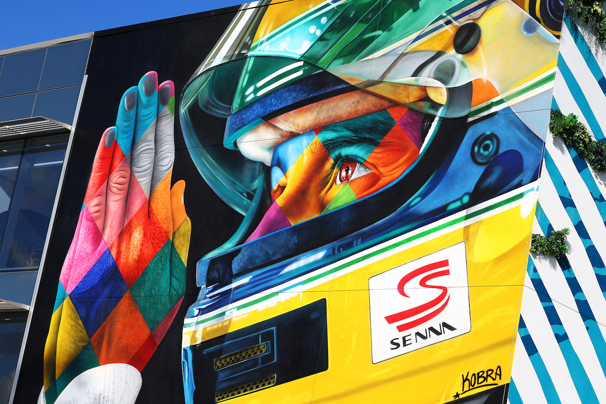 A colorful mural of Ayrton Senna in his yellow racing helmet with his hands clasped together as if praying in front of his visor.