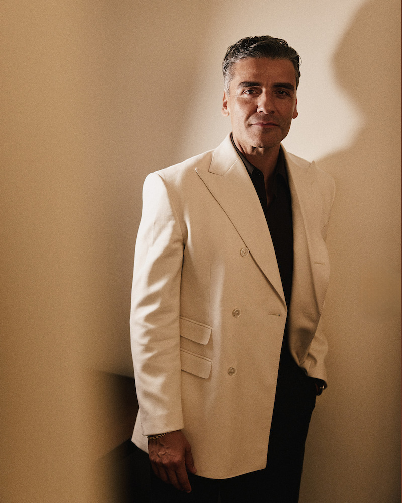 Man in a cream-colored double-breasted blazer stands indoors against a softly lit beige wall, posing with a relaxed expression and one hand in his pocket, creating an elegant and sophisticated atmosphere.