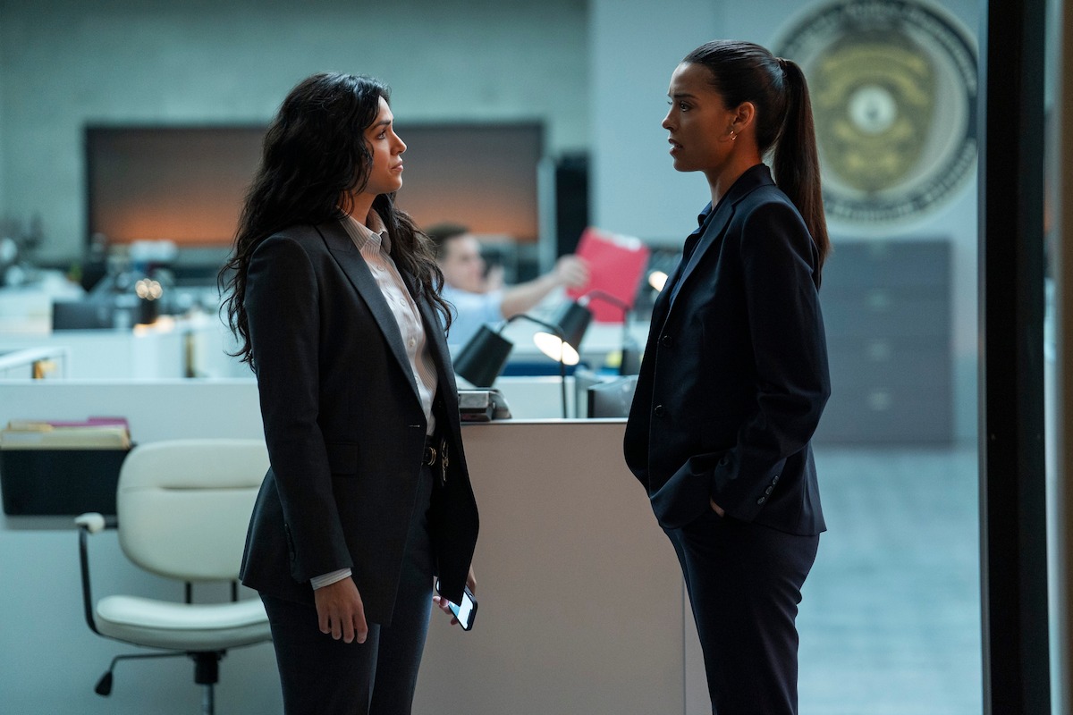 Two women in business suits stand facing each other in a modern office with desks, computers, and a seal emblem on the wall. One woman holds a phone, and an office worker is visible in the background handling files.