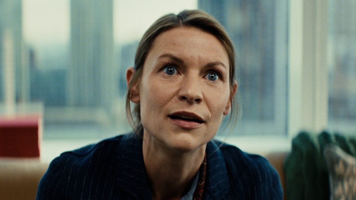 A woman with blonde hair sits indoors in an office setting, city buildings visible through large windows behind her. She looks intently forward, suggesting focus or concern. The mood is serious and tense.