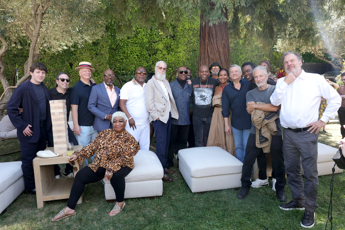 Group of diverse adults posing outdoors in a garden setting, surrounded by trees, greenery, and patio furniture, with relaxed and casual atmosphere, some seated and others standing.
