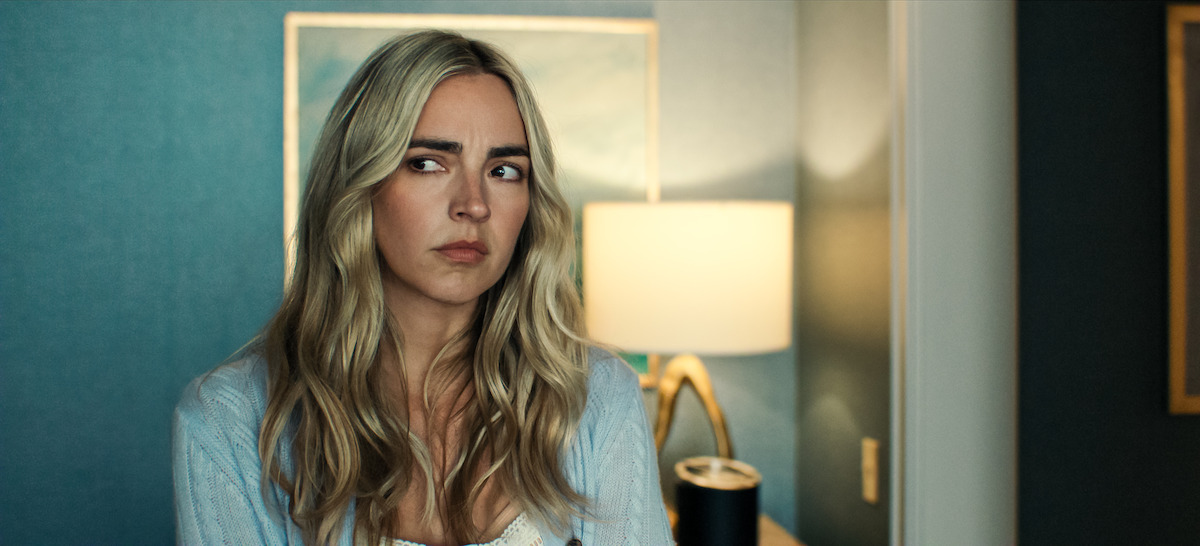 A woman with long blonde hair looks to the side with a serious expression, sitting in a room with blue walls, a painting, and a lit lamp in the background.