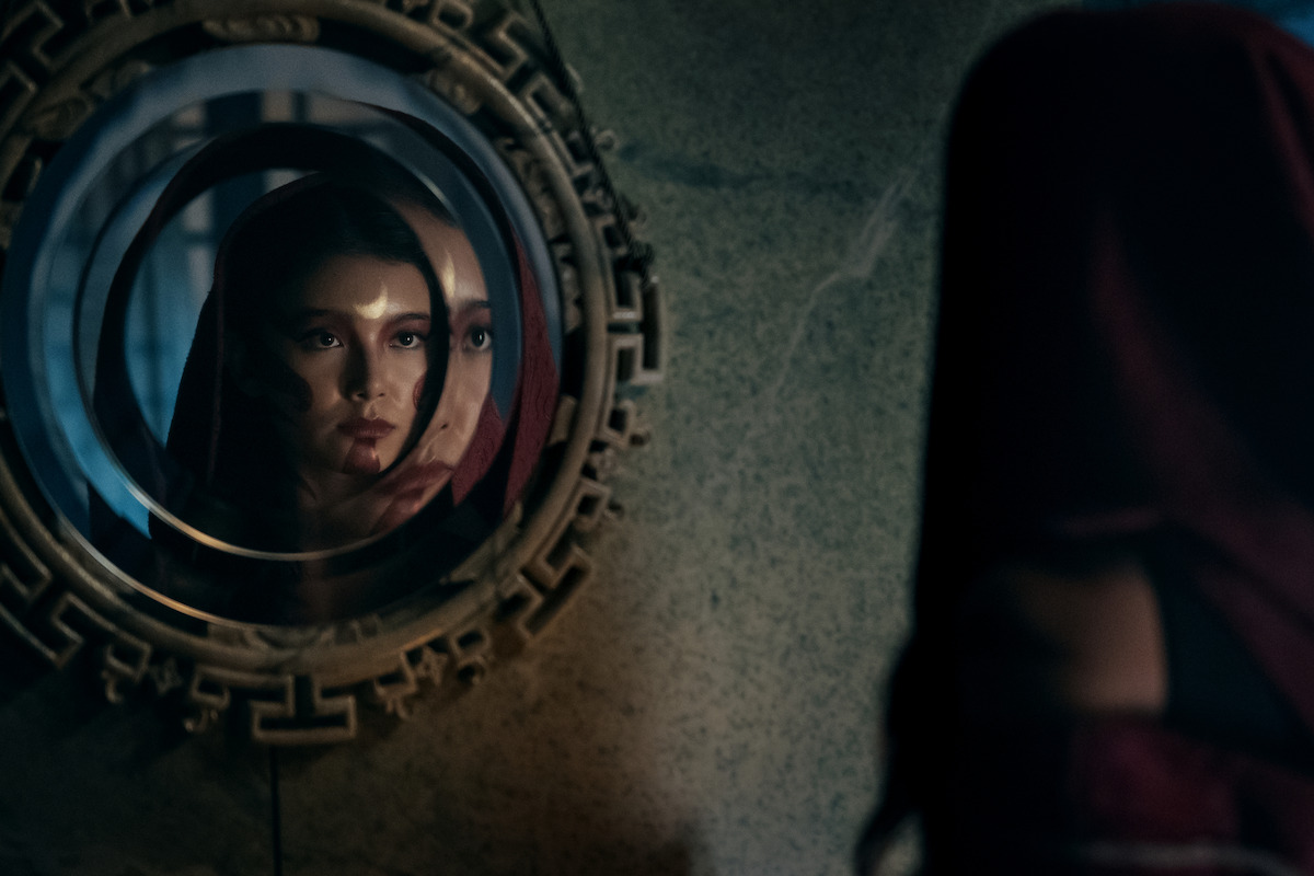 Kiawentiio as Katara in a red cloak looking into an ornate circular mirror with multiple reflections on a stone wall, creating a mysterious and dramatic atmosphere.