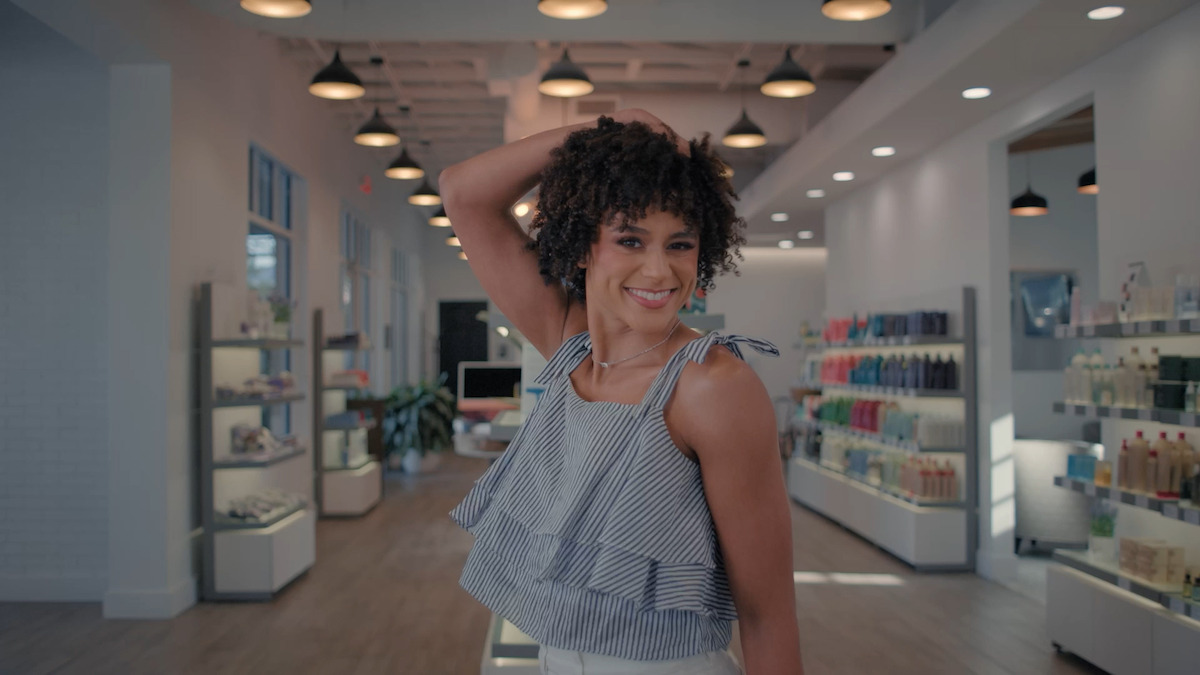 Smiling woman with curly hair posing in a modern, well-lit beauty store with shelves of products and bright overhead lights.