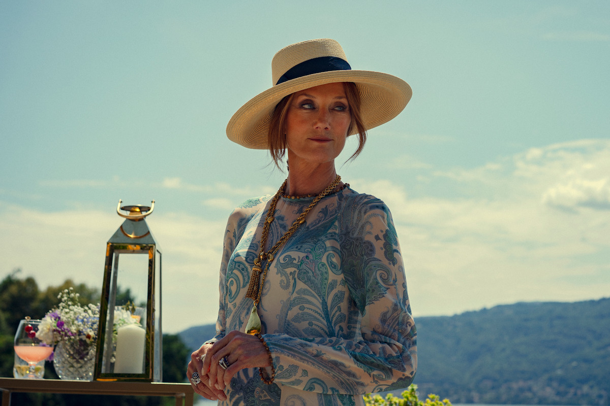 Woman in a patterned dress and wide-brim hat stands outdoors beside a decorative lantern and flowers, with hills and a blue sky in the background, suggesting a stylish outdoor gathering or event.