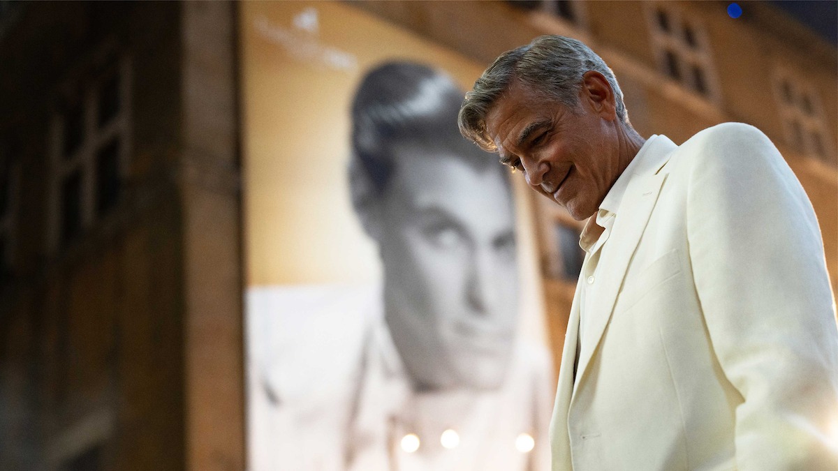 George Clooney in a white suit smiles and looks down while standing in front of a large, blurry black-and-white portrait of himself as a younger man on a building wall.