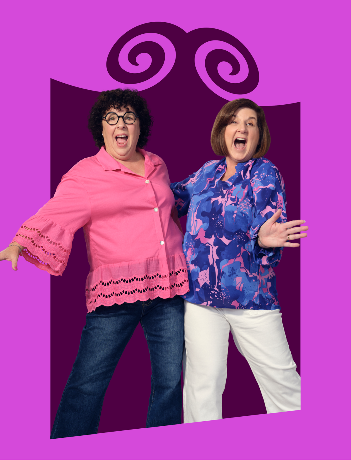Two joyful women pose together, one in a pink top and dark jeans, the other in a blue patterned shirt and white pants, standing in front of a bright purple backdrop with a whimsical decorative design.