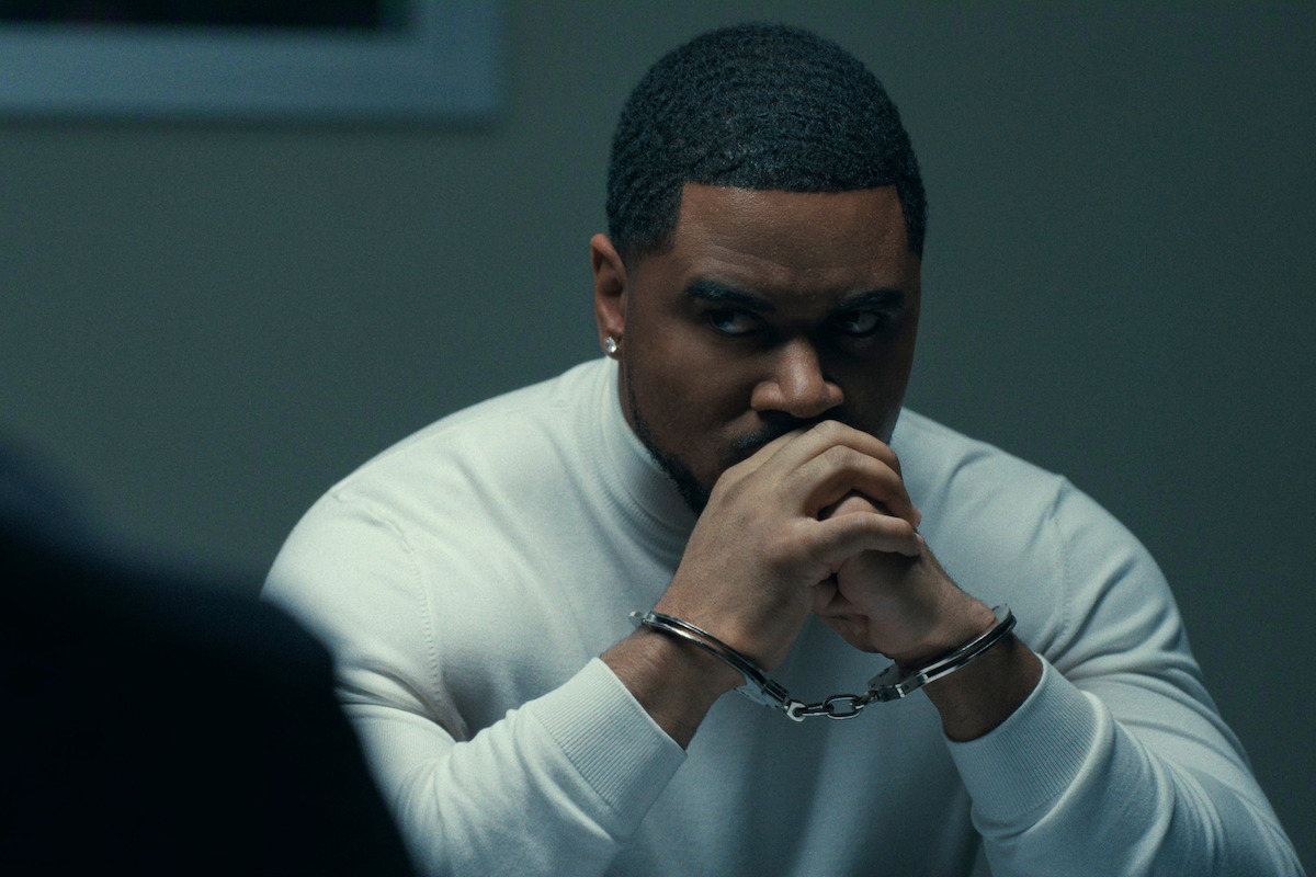A man in a white sweater sits at a table in handcuffs, looking serious and contemplative in a dimly lit interrogation room with another person partially visible across from him.