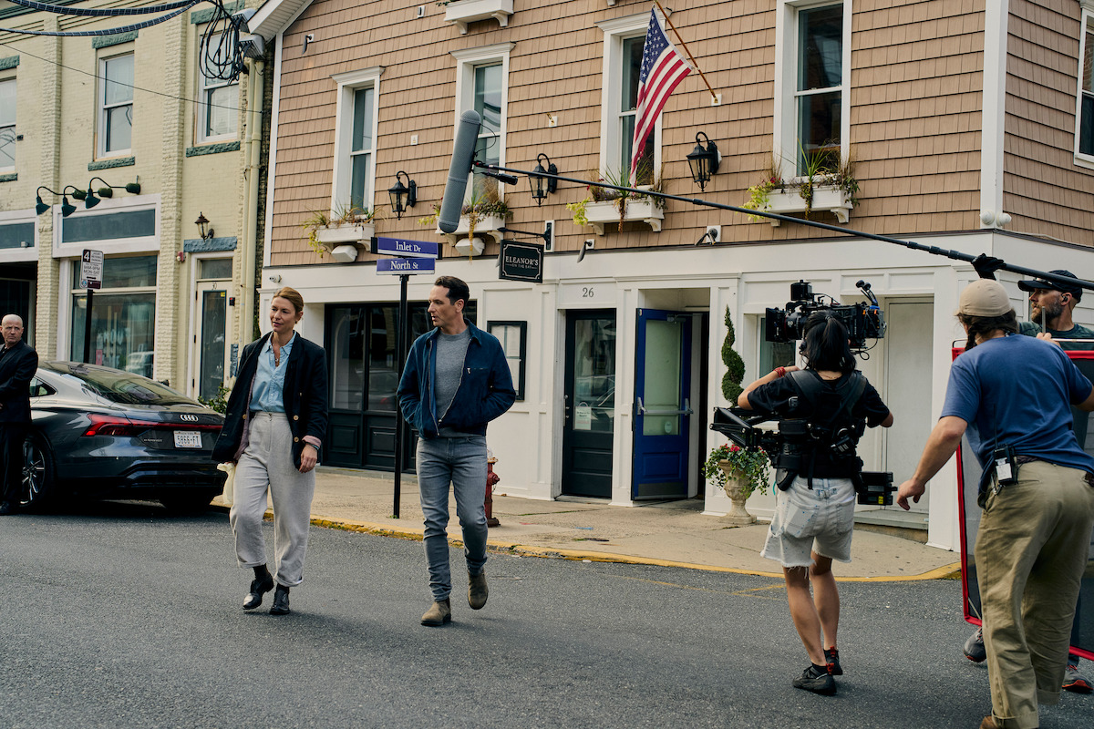 Two people are being filmed walking down a street in front of shops, with a film crew using a camera and boom mic. The environment is a small town, the mood is casual, and it appears to be a movie or TV show set in progress.