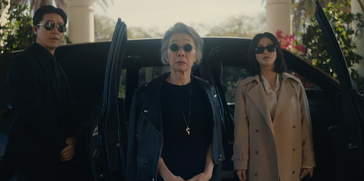 Three people wearing sunglasses stand in front of a black car with open doors, outdoors near palm trees and pillars, dressed in dark or neutral clothing, appearing serious and confident.