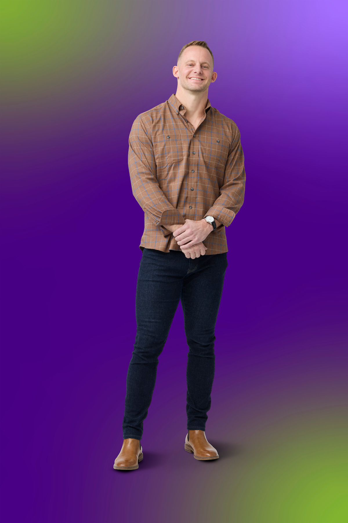 A man stands smiling with hands clasped, wearing a brown checked shirt, dark jeans, and brown shoes against a vivid purple gradient background with green accents. Studio lighting.