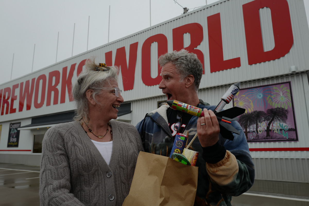 Harper Steele and Will Ferrell smile outside a store holding a brown paper bag of fireworks. Uh oh!