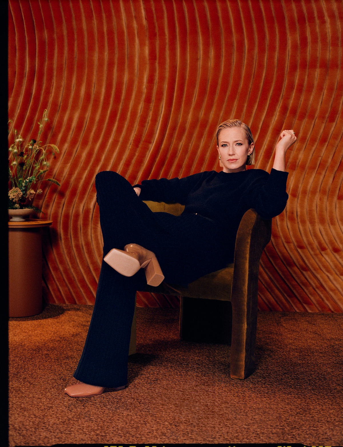 Carrie Coon wears a black ensemble and and chunky boots and sits in a chair in a burnt orange room