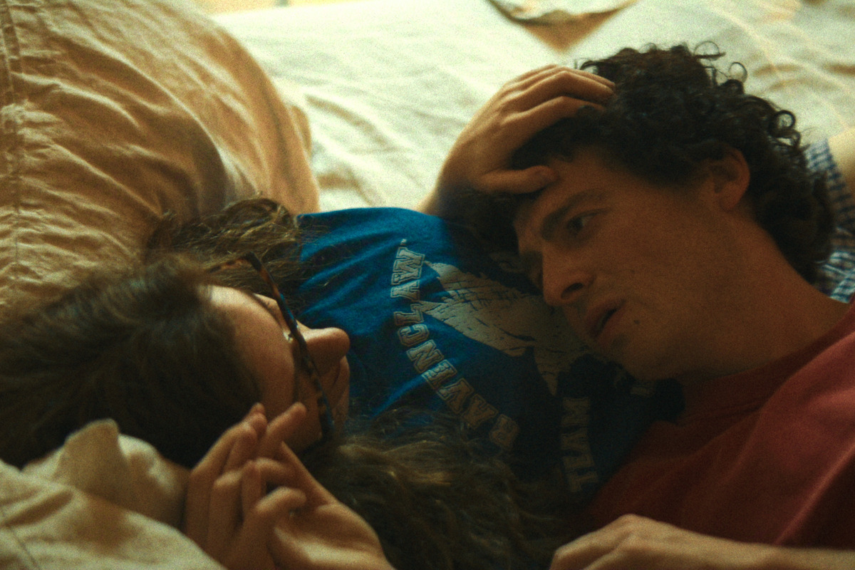 Two people lying closely together on a bed in a cozy, dimly lit room, gazing at each other and sharing an intimate moment, creating a sense of warmth and connection.