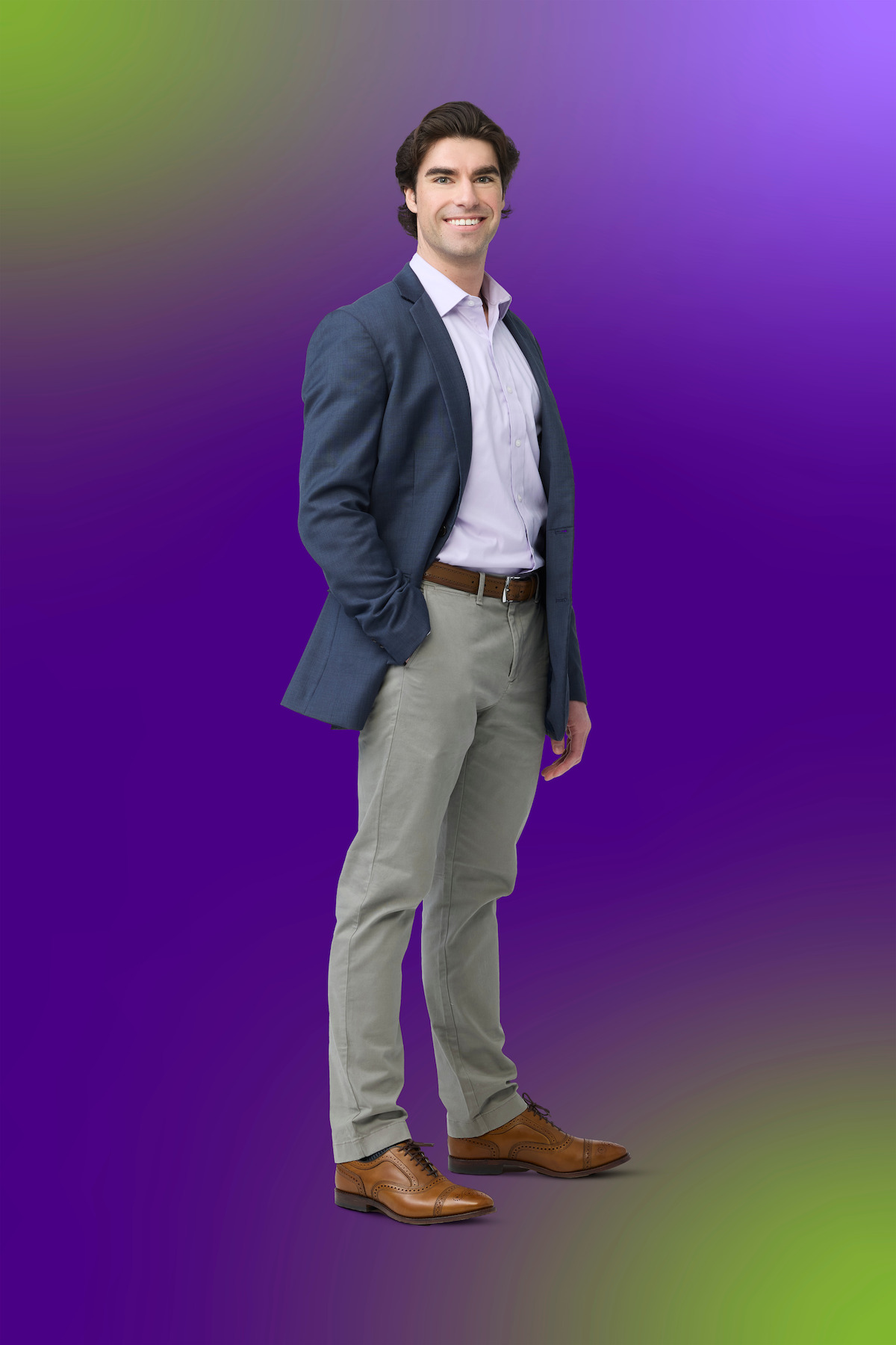 Man wearing a blue blazer, white shirt, beige pants, and brown shoes stands smiling with one hand in his pocket against a colorful gradient purple and green background.Man wearing a blue blazer, white shirt, beige pants, and brown shoes stands smiling with one hand in his pocket against a colorful gradient purple and green background.