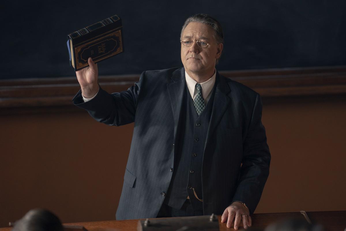 A serious man in a dark suit stands at a desk in a classroom, holding up a book in one hand. He faces the audience, with a blackboard and wood accents in the background, suggesting a formal academic environment.