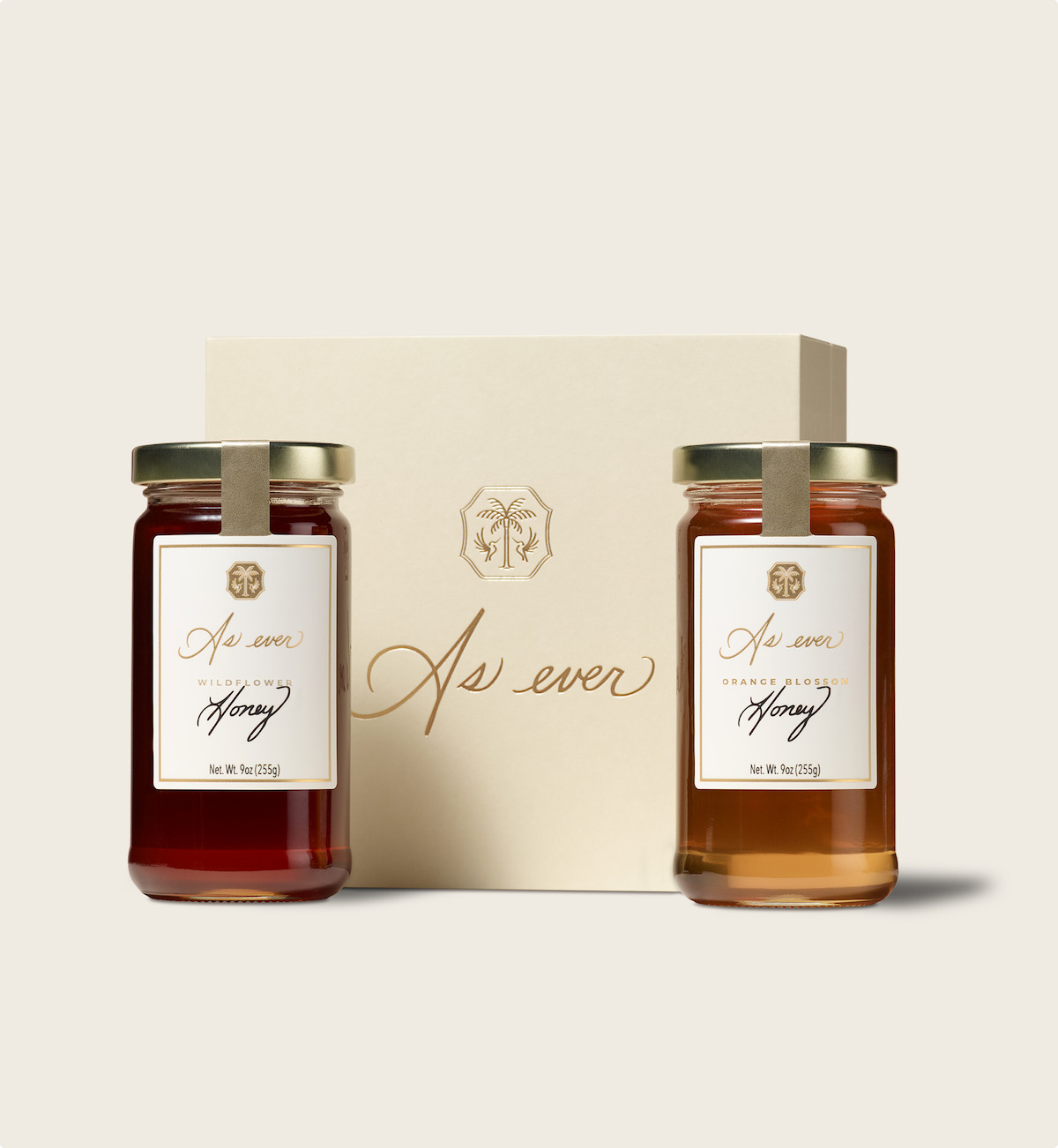 Two jars of artisanal honey, Wildflower and Orange Blossom, with elegant labels and gold lids, are placed in front of a cream-colored gift box, set against a light background, creating a refined and inviting mood.