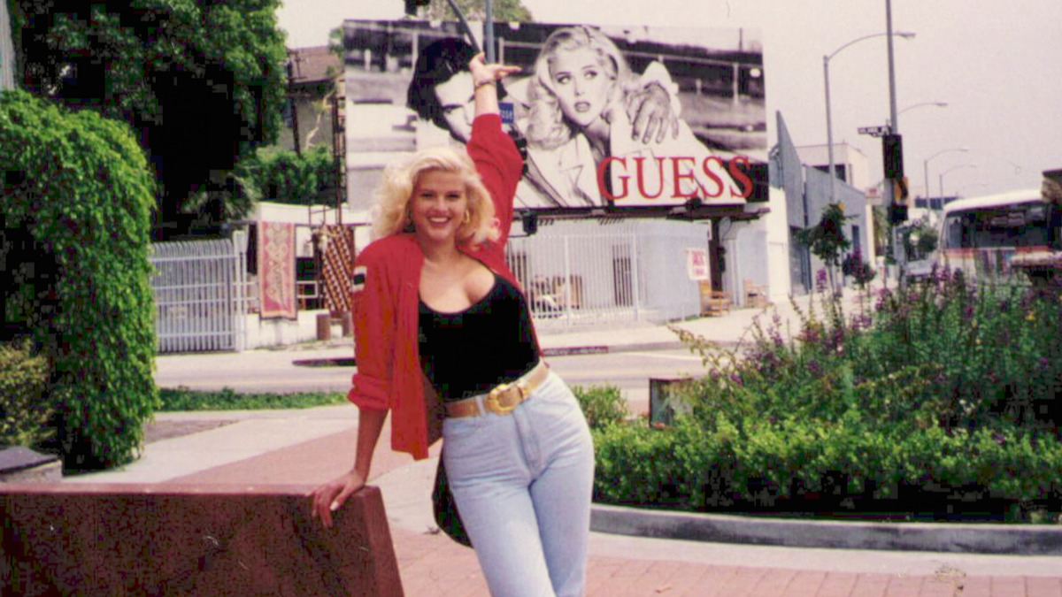 Anna Nicole Smith stands in front of a Guess billboard featuring her image. 
