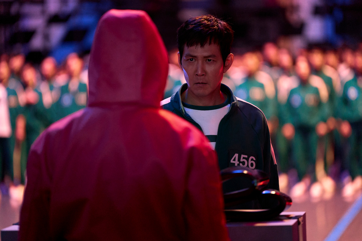 Gi-hun (Lee Jung-jae) wears a green tracksuit and stands across from a Squid Game guard in a red tracksuit.