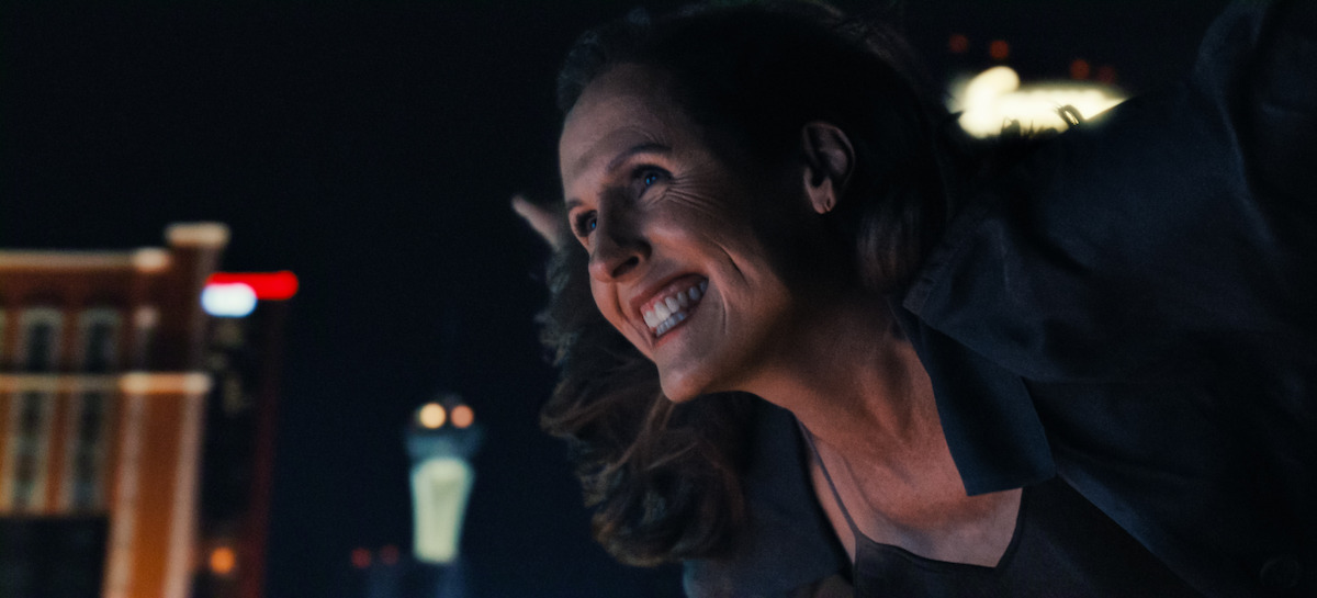Smiling woman leaning forward at night, city lights and tall buildings in the background, urban environment with a joyful expression.
