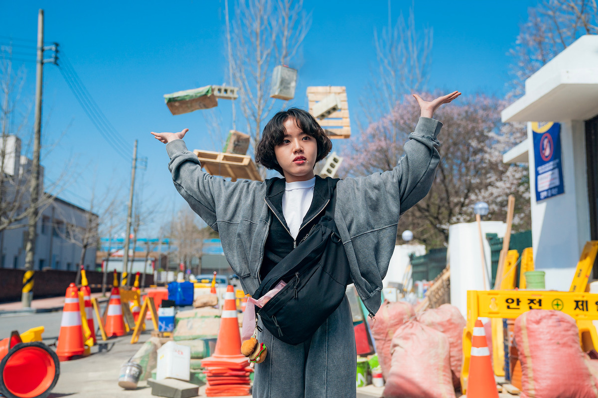 A person stands confidently at a sunny construction site with arms raised as bricks float midair, surrounded by cones and warning signs. The mood is surreal and lively, blending urban environment and playful action.