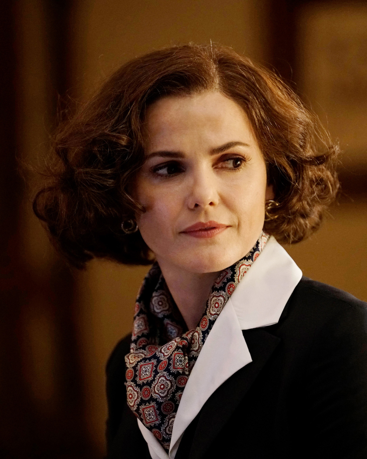 Woman with short brown curly hair, wearing a black blazer with a white lapel and a patterned scarf, indoors with warm lighting, looking to the side with a serious expression.