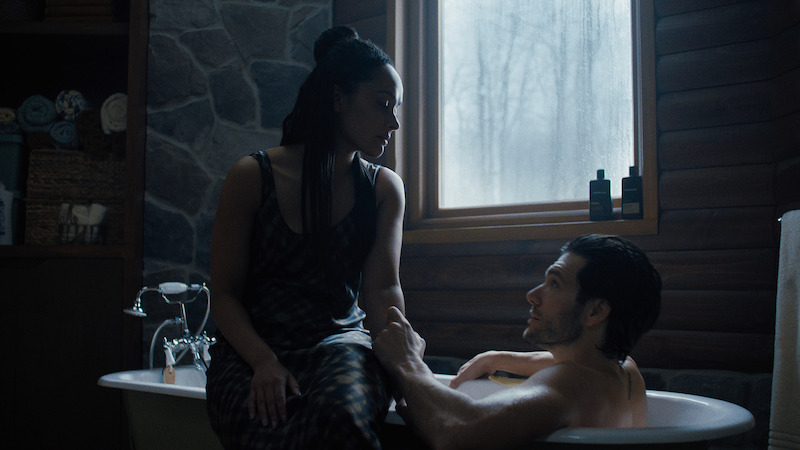 A woman in a dress sits on the edge of a bathtub beside a shirtless man in a rustic bathroom with stone and wood walls, soft light coming through a window, creating an intimate, cozy atmosphere.