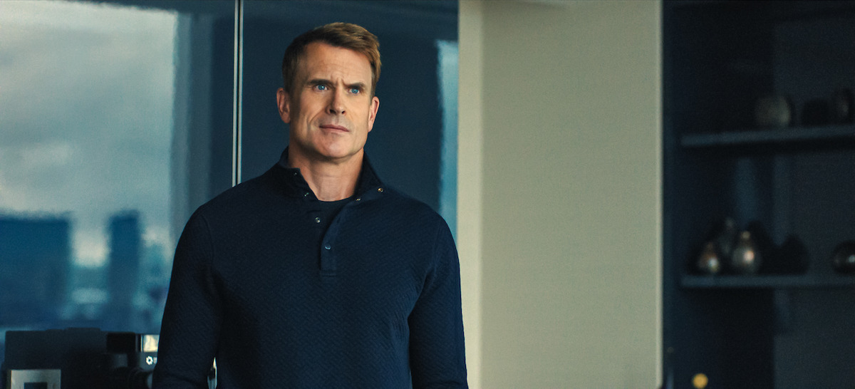 A man in a dark blue long-sleeve shirt stands indoors in a modern office with large windows and shelves holding decorative vases behind him.