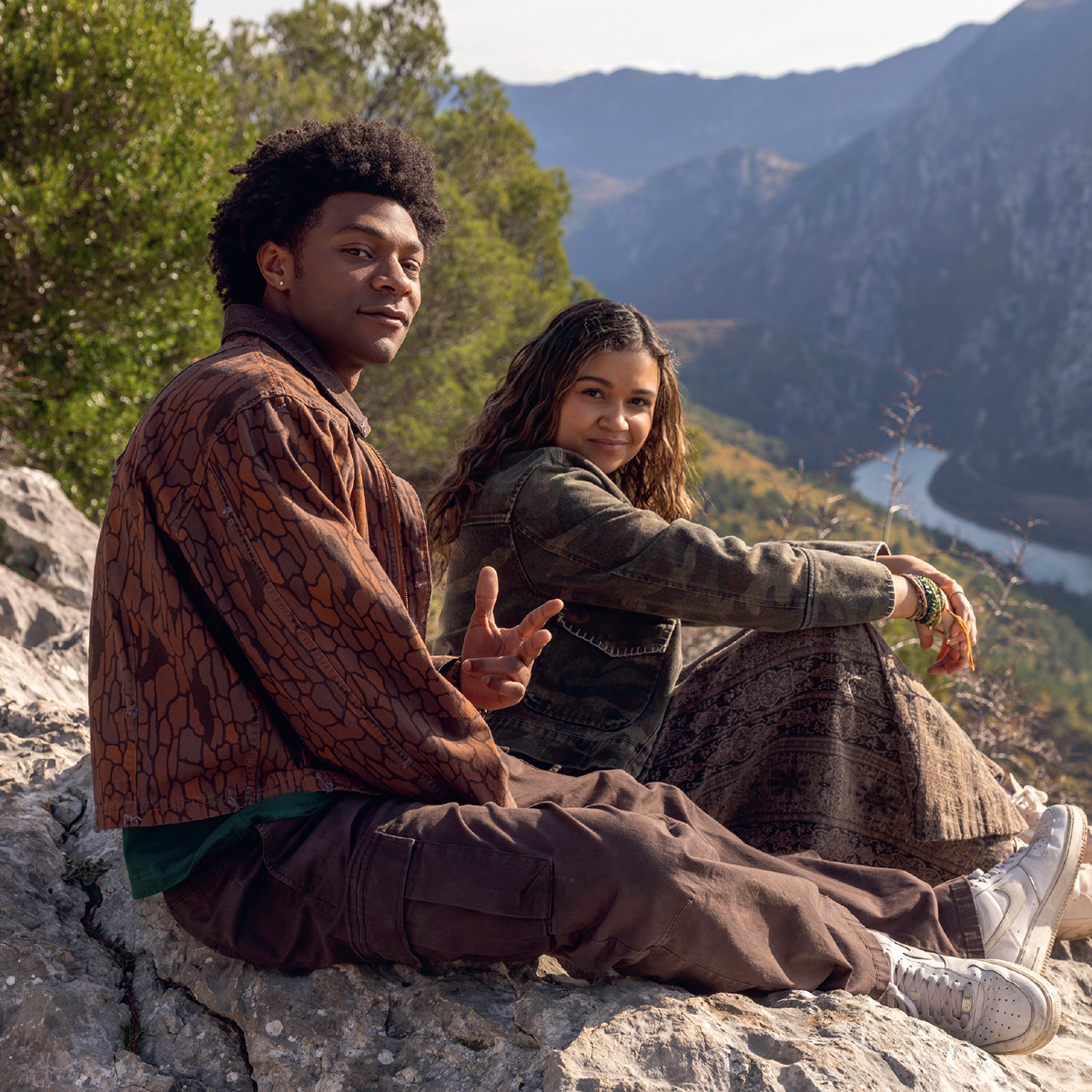 Two people sitting on rocky ground outdoors, surrounded by trees and mountains, overlooking a winding river below. Both wear casual clothes and appear relaxed, enjoying the natural landscape and scenic mountain view.