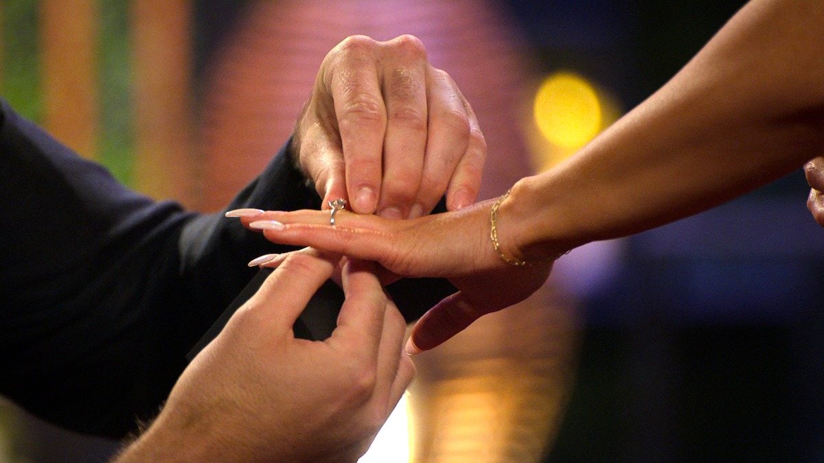 A close-up of a person placing an engagement ring on another person's finger indoors, with warm, blurred lights in the background, suggesting a romantic or special occasion setting.
