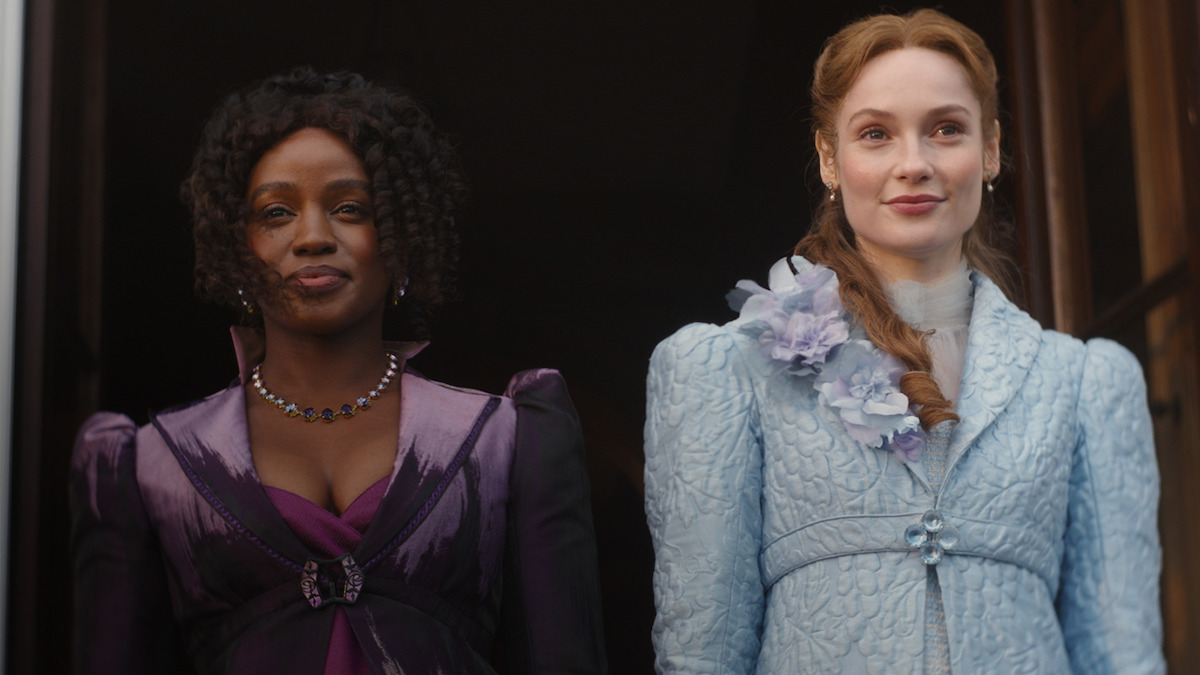 Two women in elaborate period costumes, one in purple and one in light blue with floral details, stand side by side in front of a dark background, looking forward with composed expressions in a historical setting.