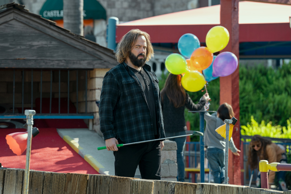 Bearded man holding mini golf club at an outdoor mini golf course, standing near a small wooden structure; in the background, children hold colorful balloons and play under a canopy on a sunny day.