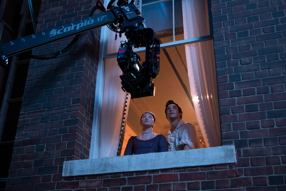 Two actors in period costumes look out a window from inside a brick building while a large camera crane films them at night, with curtains and warm interior lights in the background.