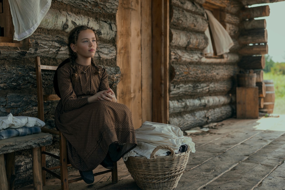 Young woman in a brown dress sits on a wooden chair beside a log cabin, with laundry in a basket at her feet and folded clothes nearby, suggesting a rural, historical setting with natural light and rustic surroundings.