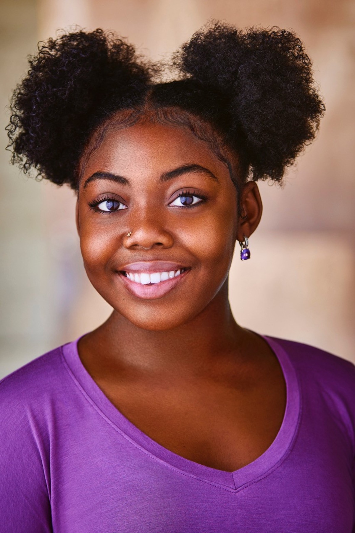 Smiling young woman with natural curly puffs hairstyle, wearing a purple top and matching earrings, standing indoors with a soft neutral background, looking directly at the camera.