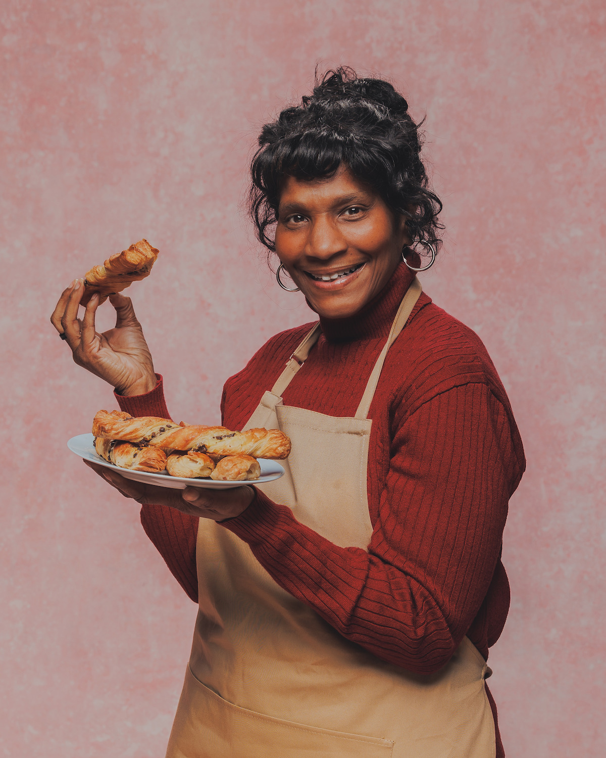 Woman wearing a tan apron and red sweater holding a plate of pastries and smiling, standing in front of a pink textured background, studio lighting.