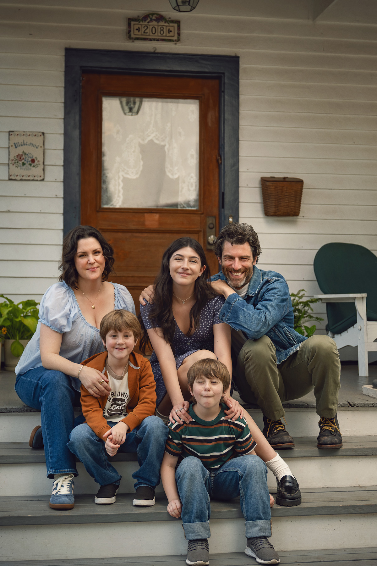 A family of five, including two adults, a teen girl, and two young boys, sits together smiling on the front porch steps of a house with white siding, plants, and a wooden door, suggesting a warm, casual outdoor setting.