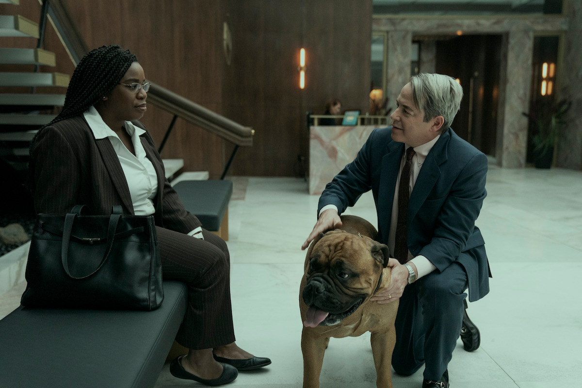 Uzo Aduba as Edie sits in a lobby as Matthew Broderick as Richard Sackler kneels near by petting a dog in Season 1 of ‘Painkiller.’