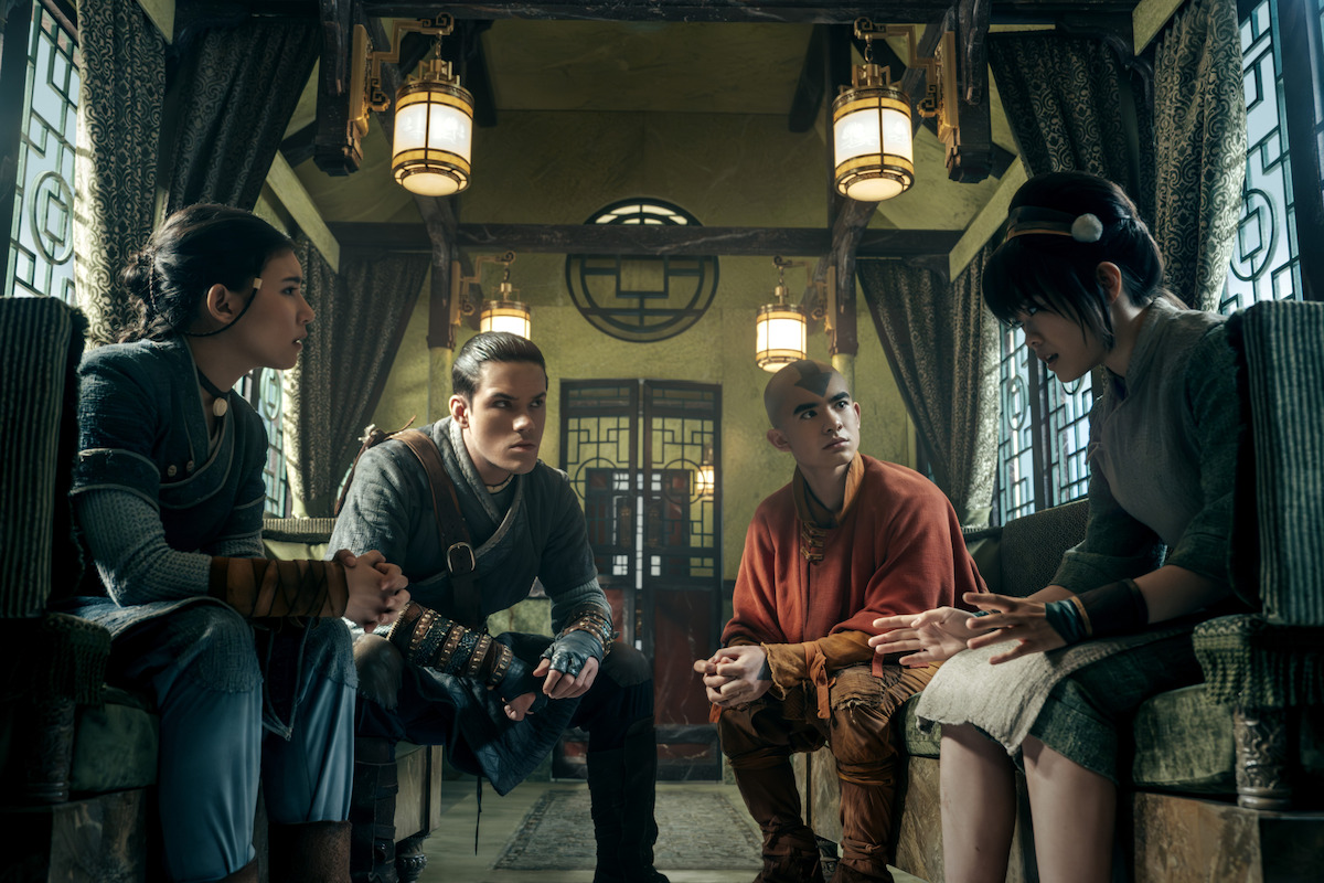 Four young people in detailed, fantasy-style clothing sit and talk seriously in an ornately decorated room with wooden beams, hanging lanterns, and patterned windows, suggesting a scene from a historical or adventure story.