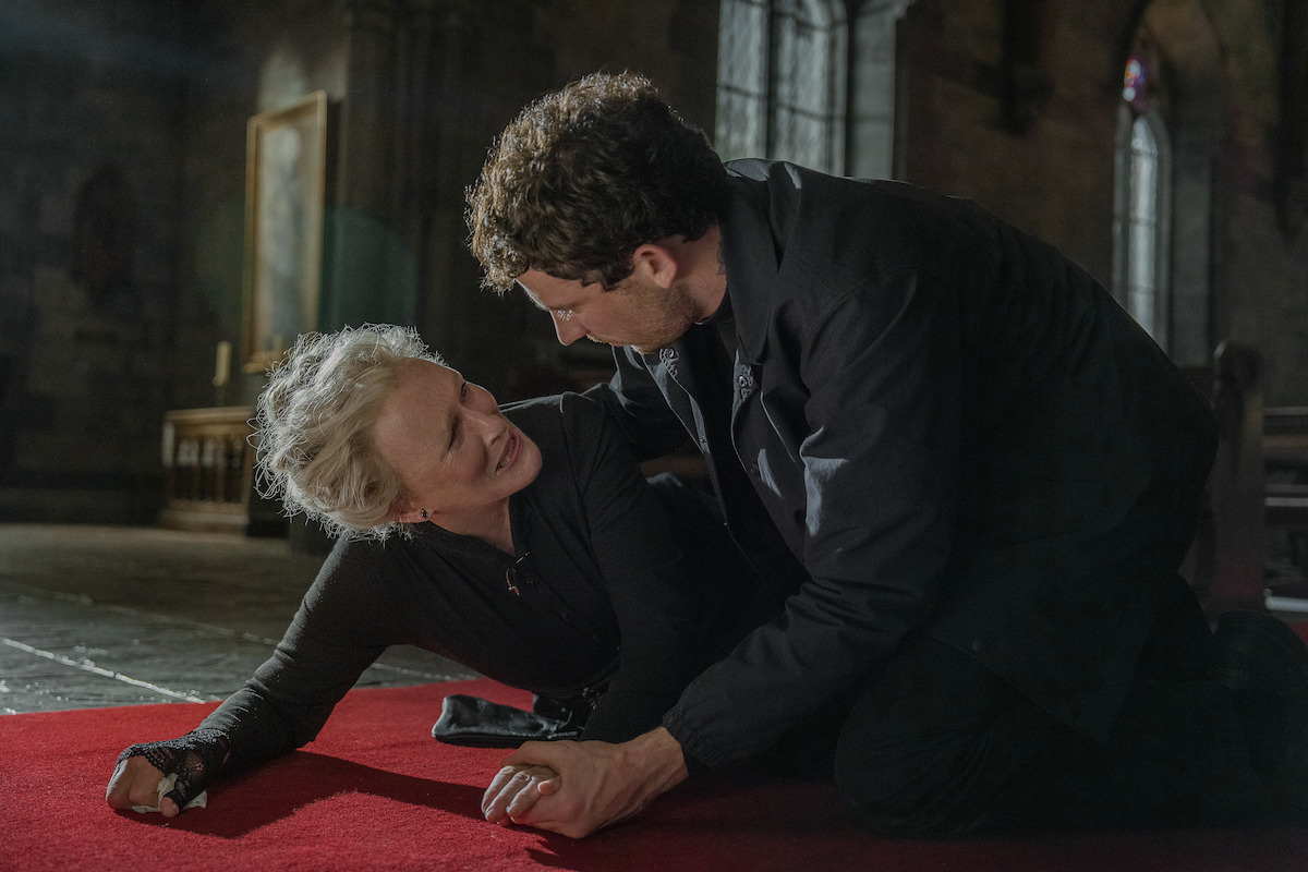 An older woman on the floor of a dimly lit church looks up in distress at a man leaning over her. The mood is tense and dramatic, with emotional intensity and gothic atmosphere in the historic setting.