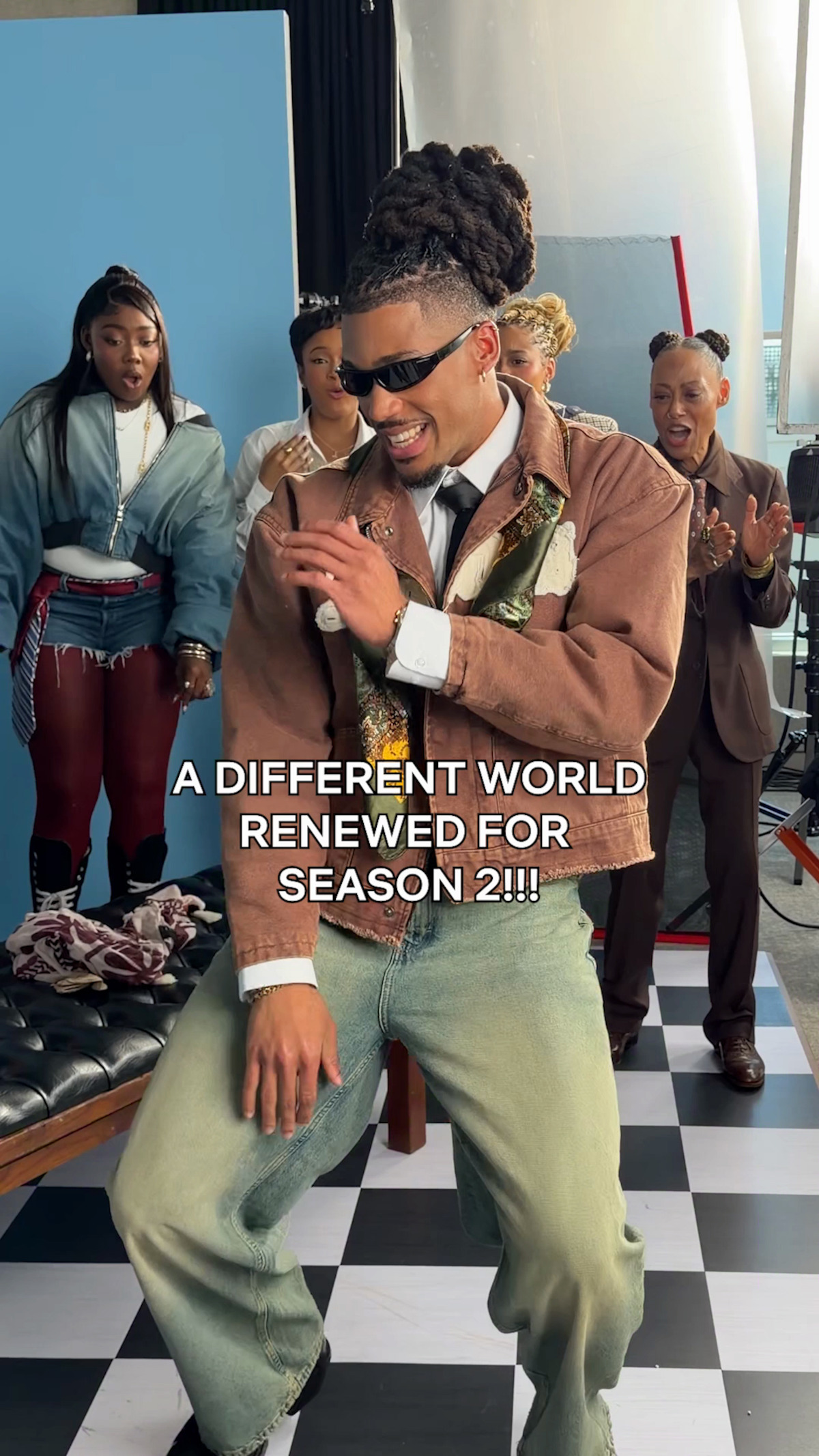 Man dancing joyfully in retro-style outfit, wearing sunglasses, with three people in colorful vintage clothing behind him on a black and white checkered floor, set in a bright studio environment. Text: A Different World renewed for season 2.