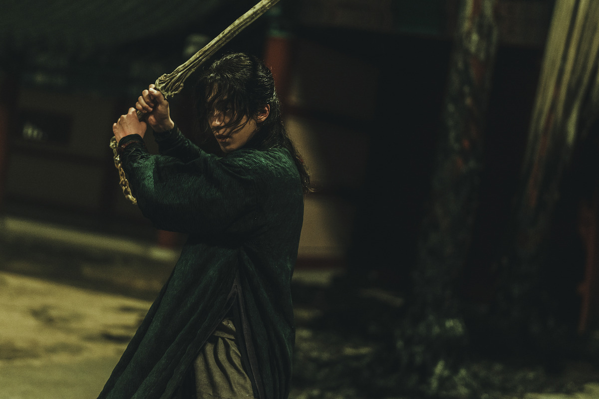 Person in traditional clothing holding a sword in a dramatic pose, poised for action in a dimly lit, historic outdoor setting, possibly during nighttime.
