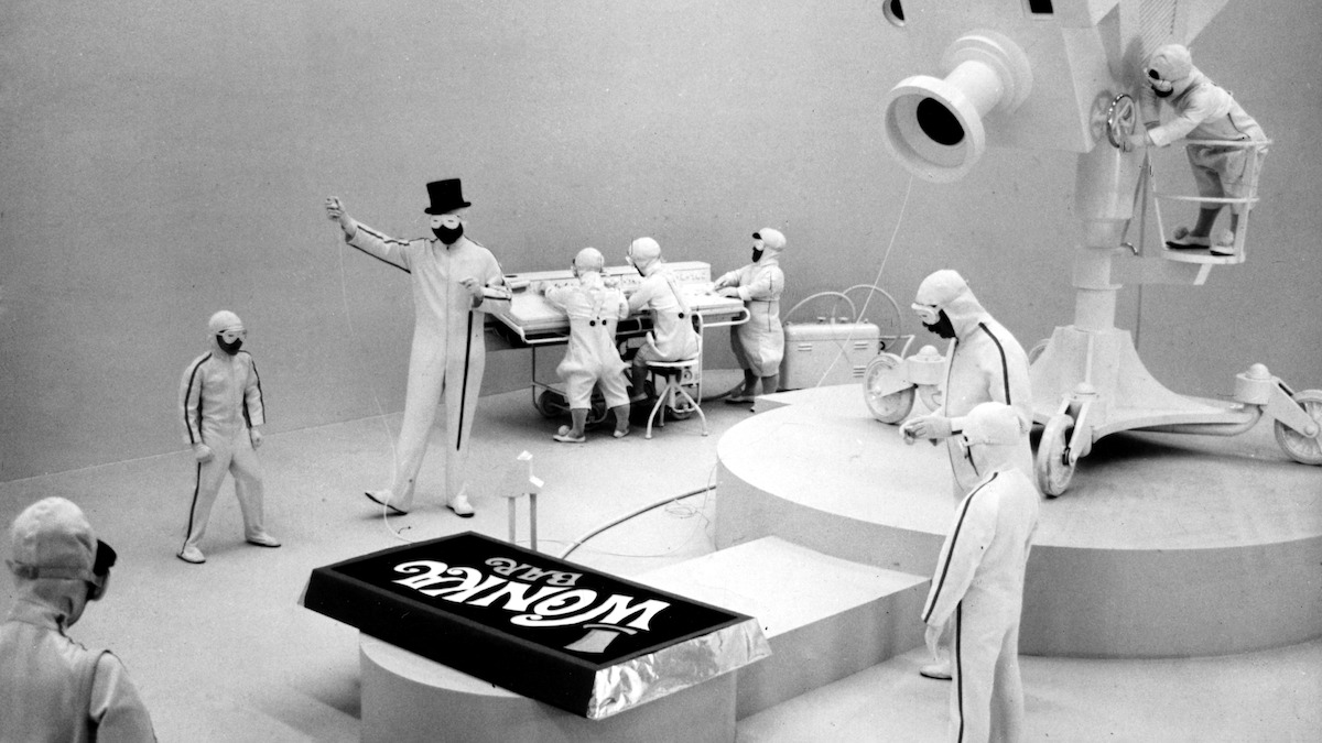 A man in a top hat and workers in clean suits stand in a sterile, futuristic factory with large machinery, examining a giant Wonka chocolate bar on a platform.