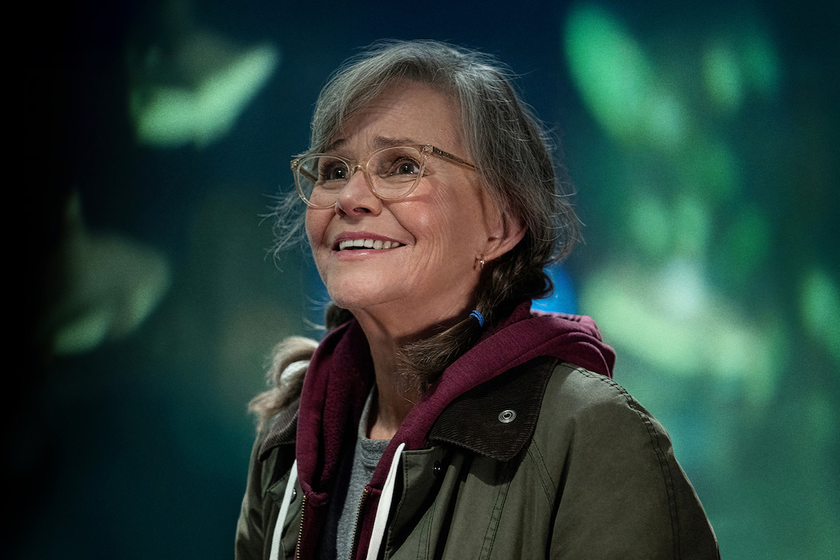 Sally Field as Tova, smiling, wearing a jacket and hoodie, standing indoors with a blurred aquarium or underwater scene in the background.