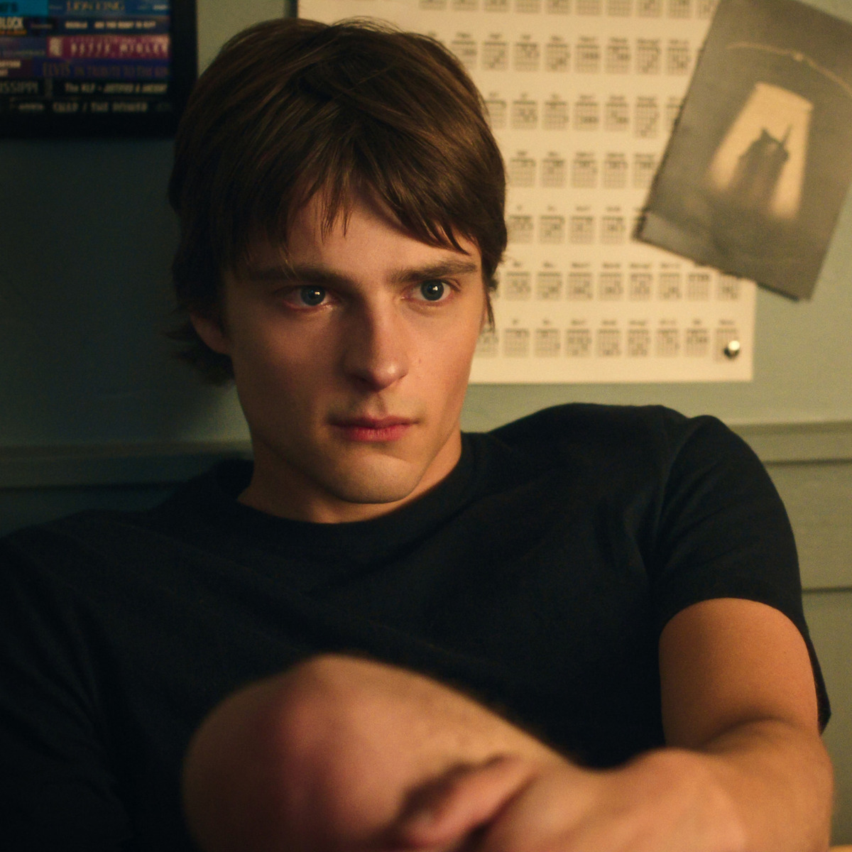Young man with brown hair sits indoors against a wall with posters and a calendar, looking thoughtful or pensive. The setting appears to be a bedroom or dorm room with soft lighting and personal decorations in the background.