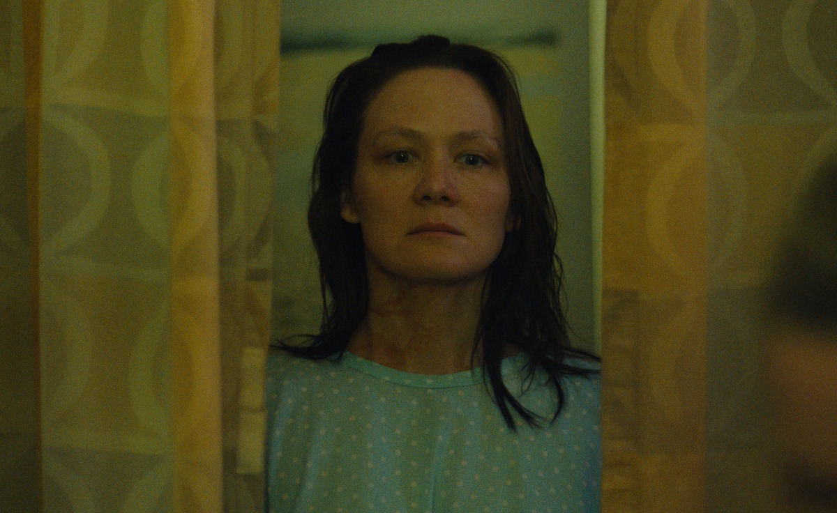 A woman with wet hair and a hospital gown stands between yellow curtains, looking serious in a dimly lit room.