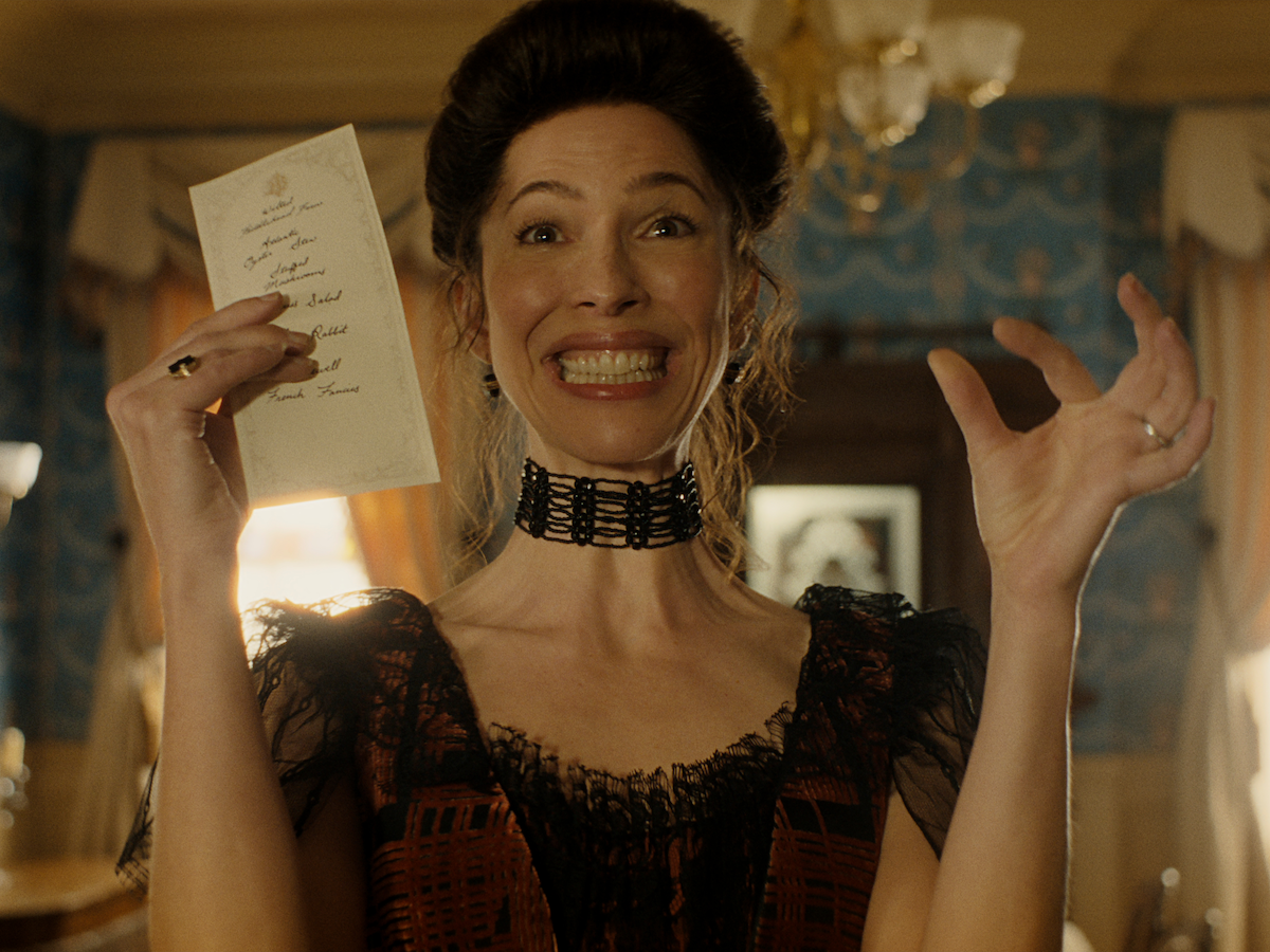 Woman in Victorian-era dress excitedly holding up a letter in a richly decorated, vintage room with ornate wallpaper, wooden furniture, and warm lighting, suggesting a historical or period drama setting.