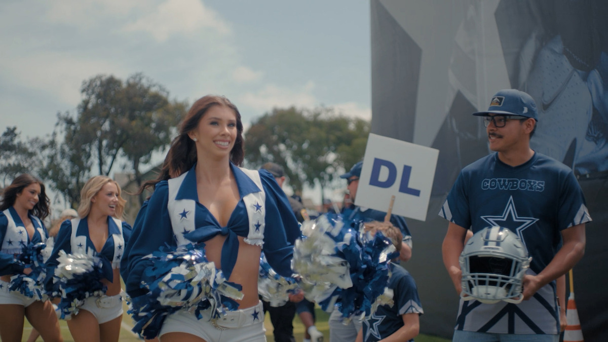 Dallas Cowboys cheerleaders in blue and white uniforms perform outdoors near a man holding a “DL” sign and a helmet, under a bright sky with trees and a team banner in the background.