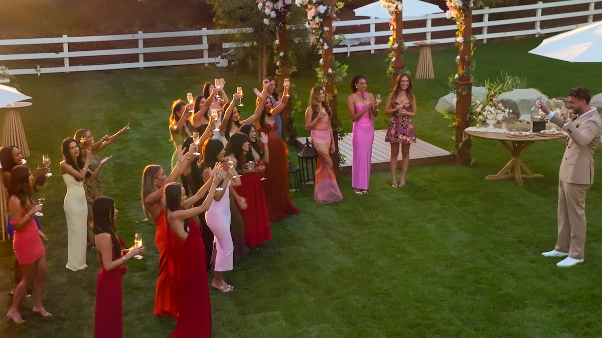 A group of women in colorful dresses raise glasses in a toast outdoors with a man in a suit under a floral arch, during a garden party or celebration on a sunny day.