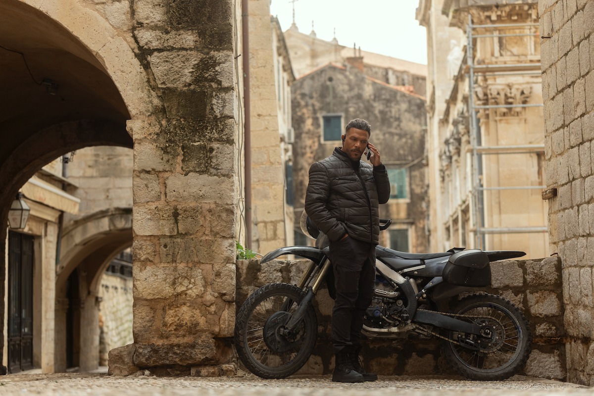 Man standing next to a black motorcycle in an old European stone alley, wearing dark clothes and talking on a phone, surrounded by ancient architecture and arches.